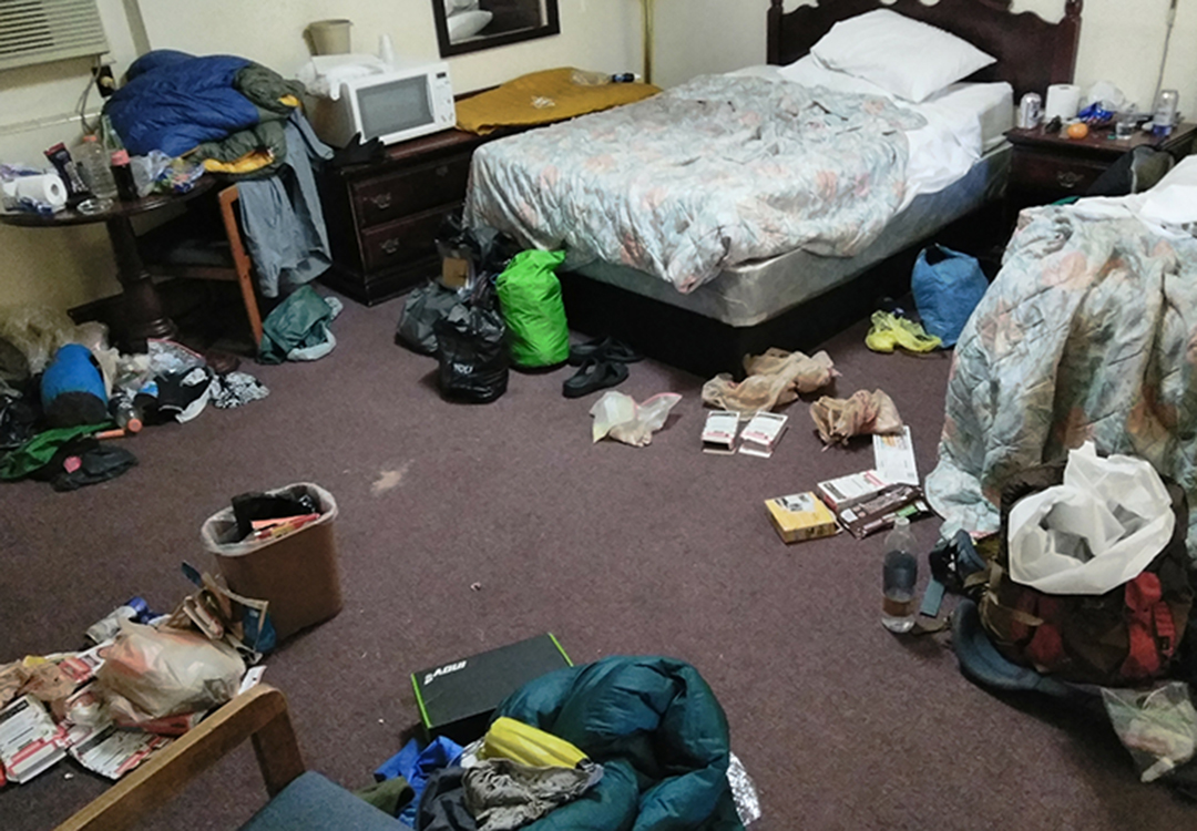 A dirty hotel room where hikers have their supplies on the floor.