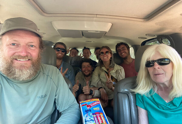 A group of backpackers smile in a car with the driver that picked them up.