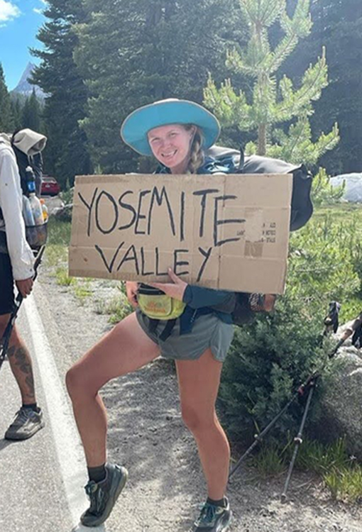 A woman with a hat on smiles as she holds up a sign that says "Yosemite Valley"