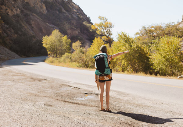 A woman stands next to a roadway with her thumbout while hitchhiking