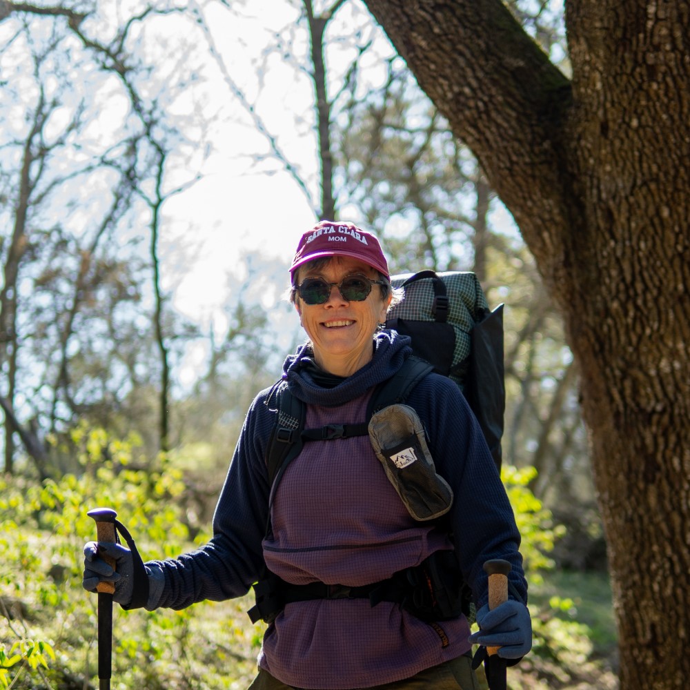 Backpacker on the trail with a ULA 2024 Catalyst attaching his bear canister to the top of his pack with a Y-strap