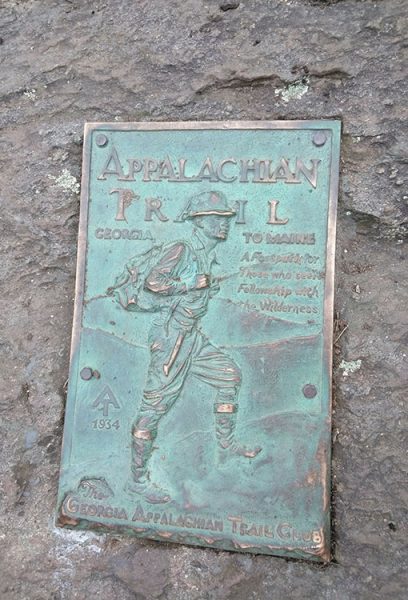 A brass Appalachian Trail plaque on a rock showing a historic hiker. The plaque says: Appalachian Trail Georgia to Maine, 1934. The Georgian Appalachian Trail Club. A forest path for those who seek fellowship with nature."