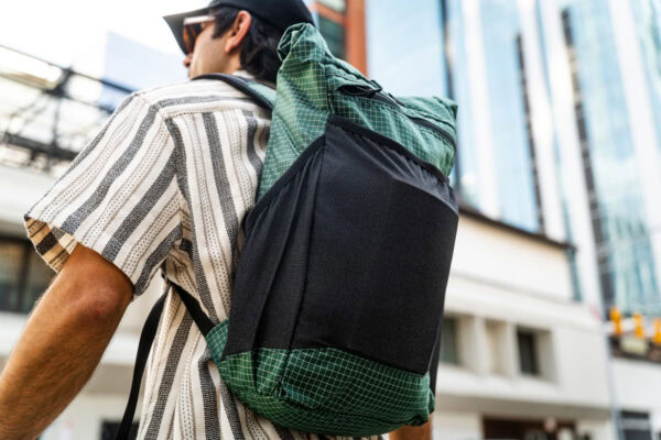 An urban explorer wears a ULA in brushed green robic fabric. There are skyscrapers behind him.