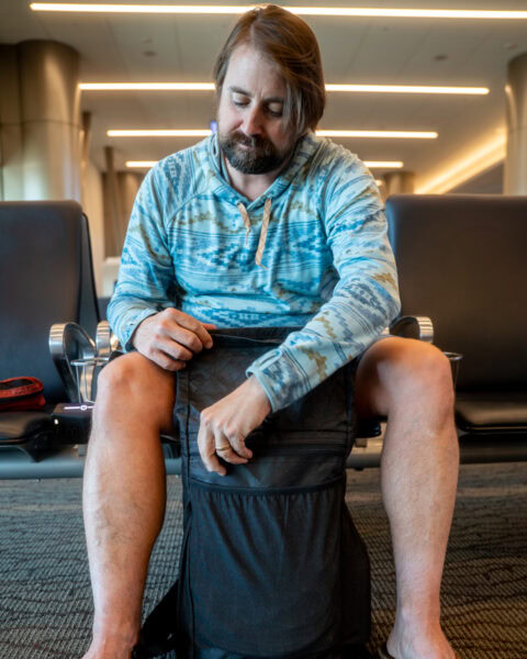A traveler in an airport unzips the external zippered pocket on the front of his ULA Ultra Node