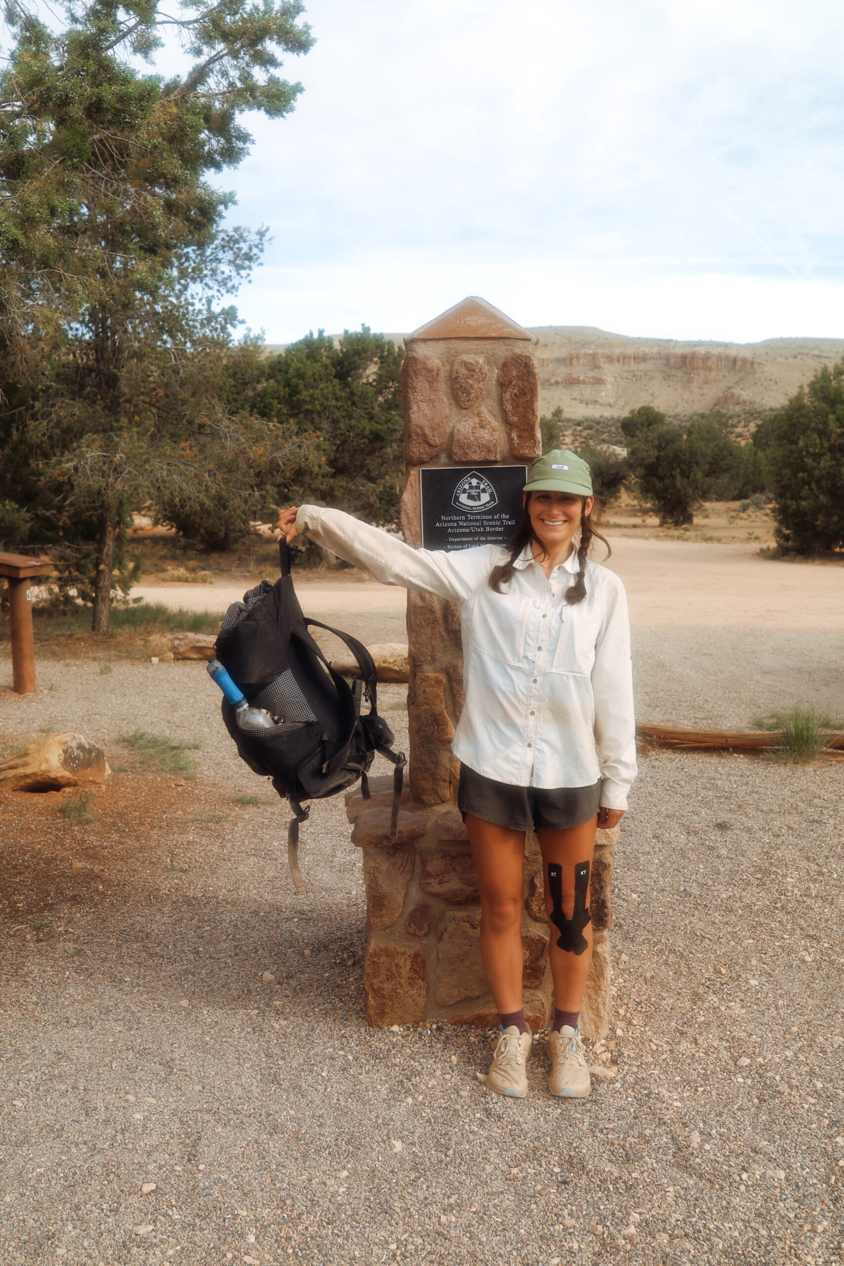 Baja on the AZT A female hiker holds up her backpack while smiling infront of the Arizona Trail terminus