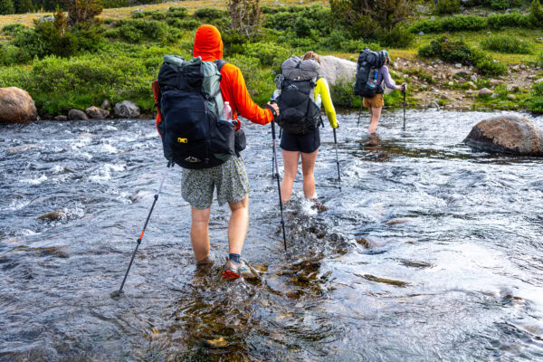 Three backpackers cross an ankle deep river in the Wyoming wilderness. They are all using ULA packs.