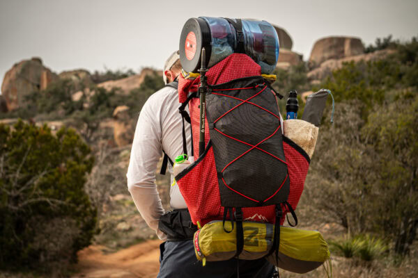 A back view of a backpacking wearing a red robic ULA pack with a tent on the bottom straps, and a bear bin under a top Y-strap.